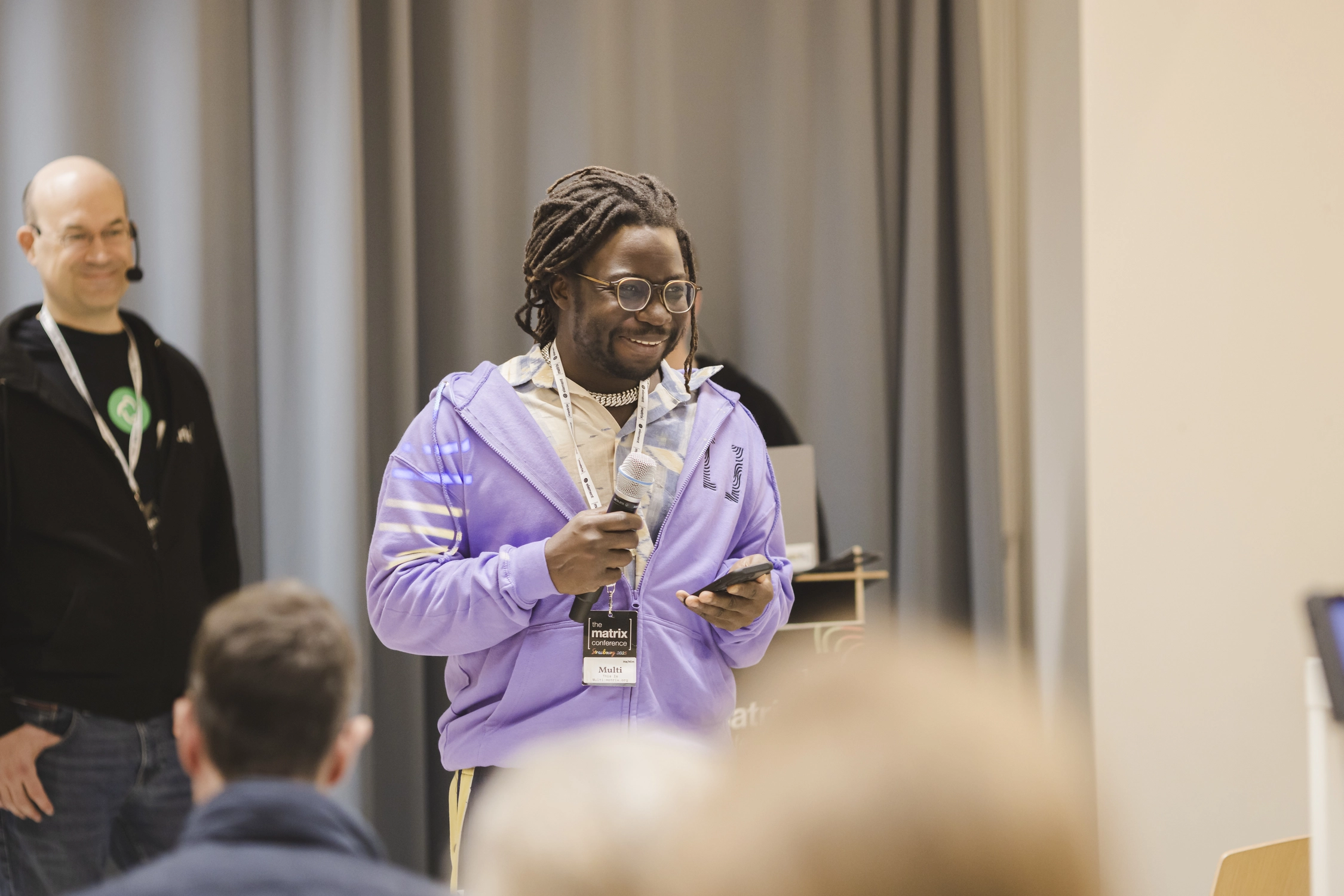 A photo of a man in a purple hoodie, holding a microphone and smiling at the audience while reading remote audience questions from his phone. In the background, the speaker is waiting to answer.